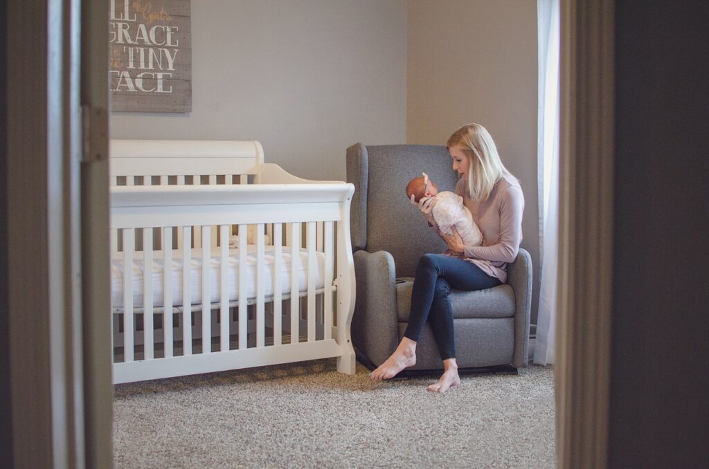 woman in nursery holding her baby, sitting on her ottoman with a white crib near by with carpet